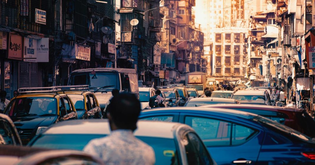 Trânsito caótico numa rua urbana densa, com automóveis parados, edifícios altos, comércio local e circulação congestionada.