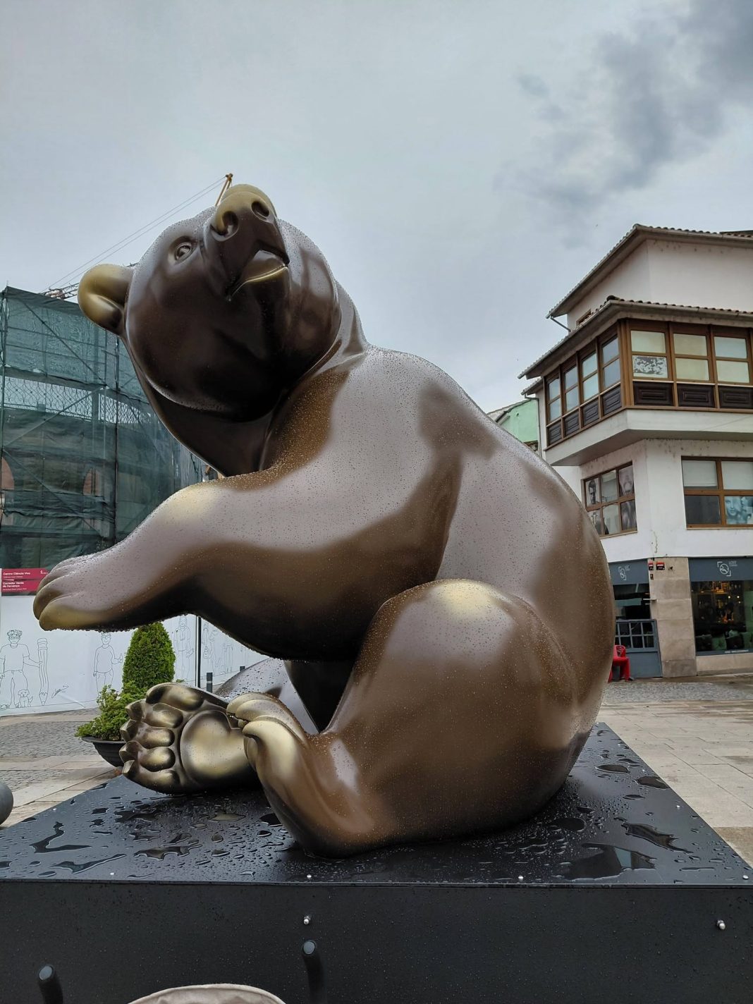 Escultura em bronze de um urso sentado, com uma pata levantada, numa praça pública de Bragança, com um edifício histórico ao fundo