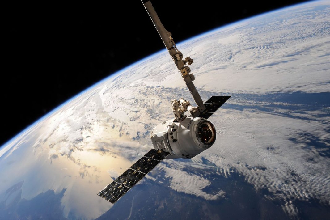 Satélite de comunicações em órbita sobre a Terra, com painéis solares abertos e a superfície coberta por nuvens.